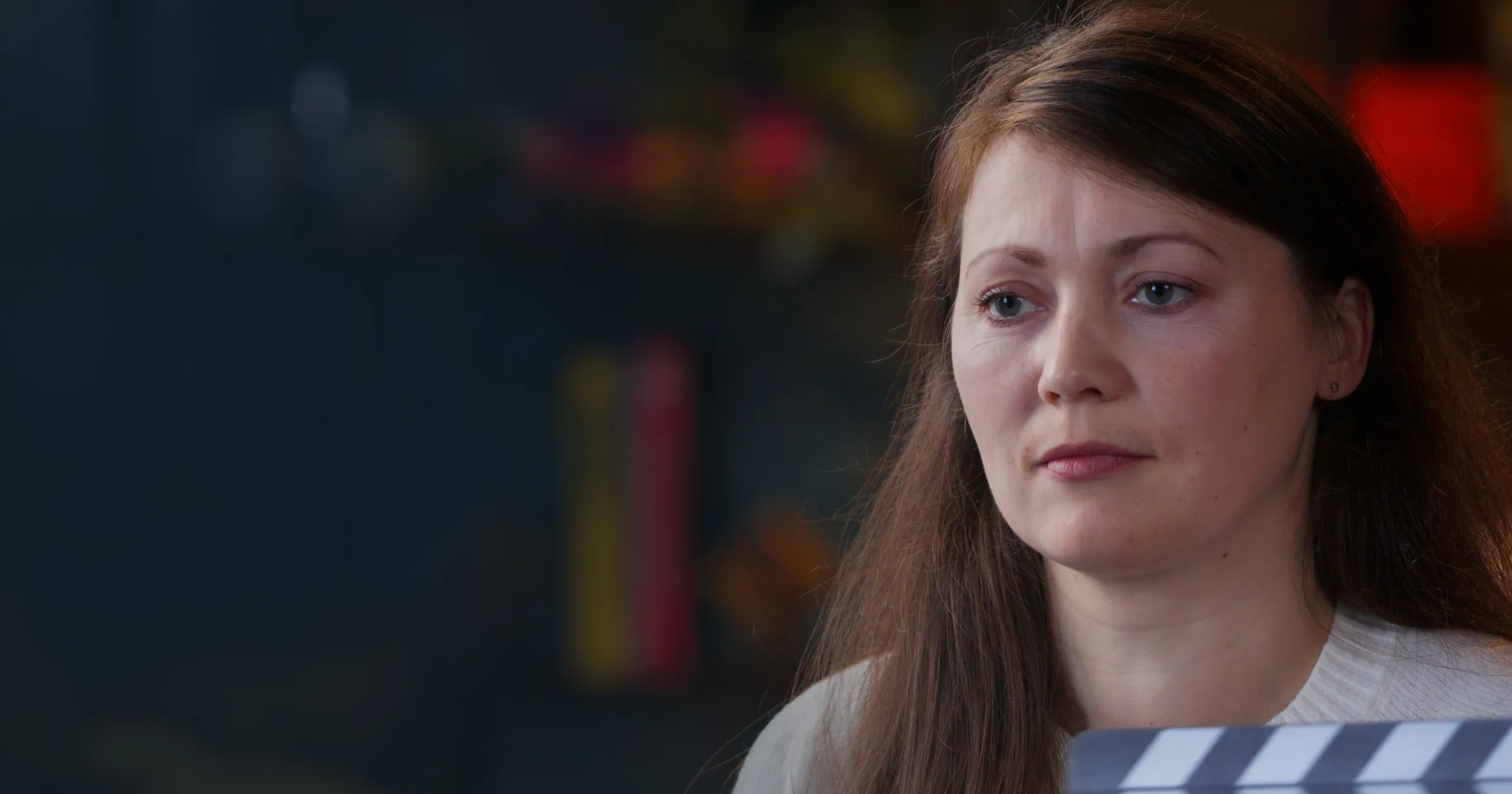A woman with long brown hair looks thoughtful, holding a movie clapperboard. The background is softly blurred with colourful, out-of-focus shelves.